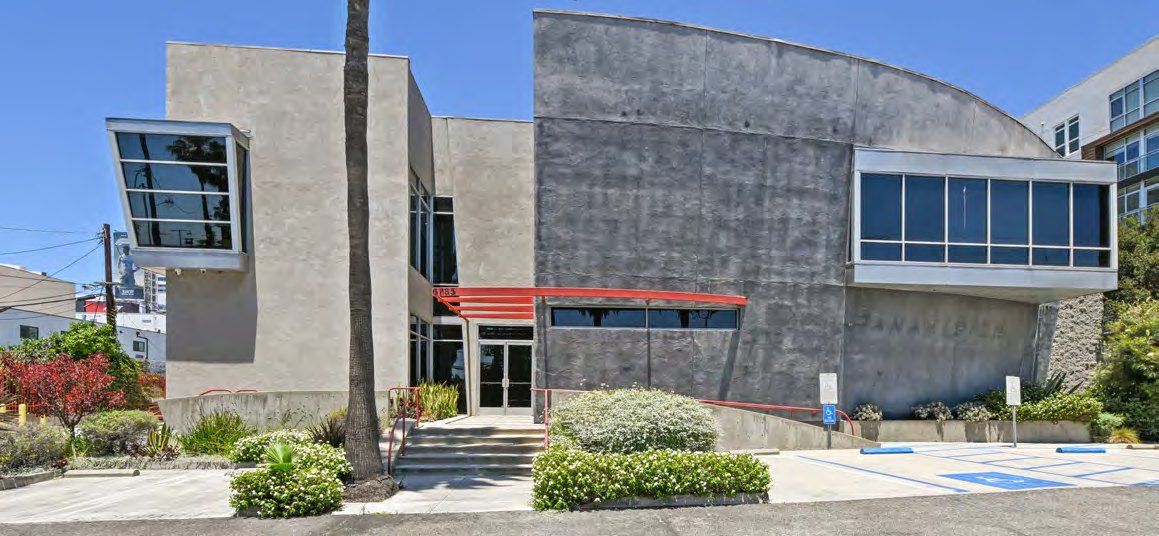 Wide exterior view of the freestanding Selma House campus from the street, showing concrete volumes and the signature orange-red awning band